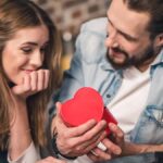Un hombre barbudo le muestra una caja en forma de corazón a su pareja, ilustrando una de las etapas de cómo se enamoran los hombres.