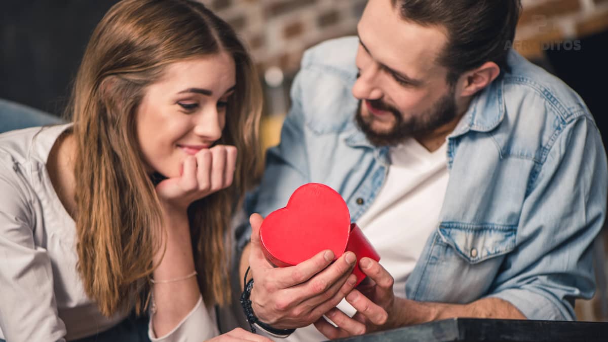 Un hombre barbudo le muestra una caja en forma de corazón a su pareja, ilustrando una de las etapas de cómo se enamoran los hombres.