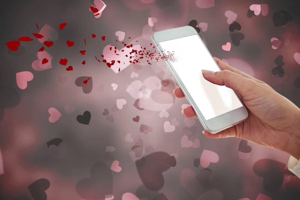 Mano sosteniendo un teléfono móvil con corazones virtuales en el fondo.